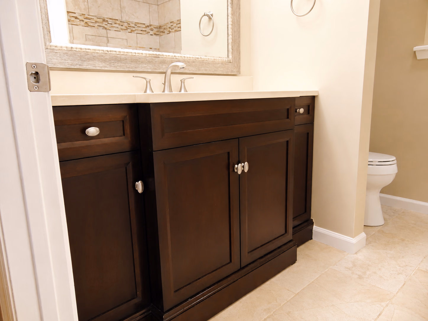 Bathroom vanity with dark wood cabinets, beige countertop, and a mirror above, toilet visible in the corner.