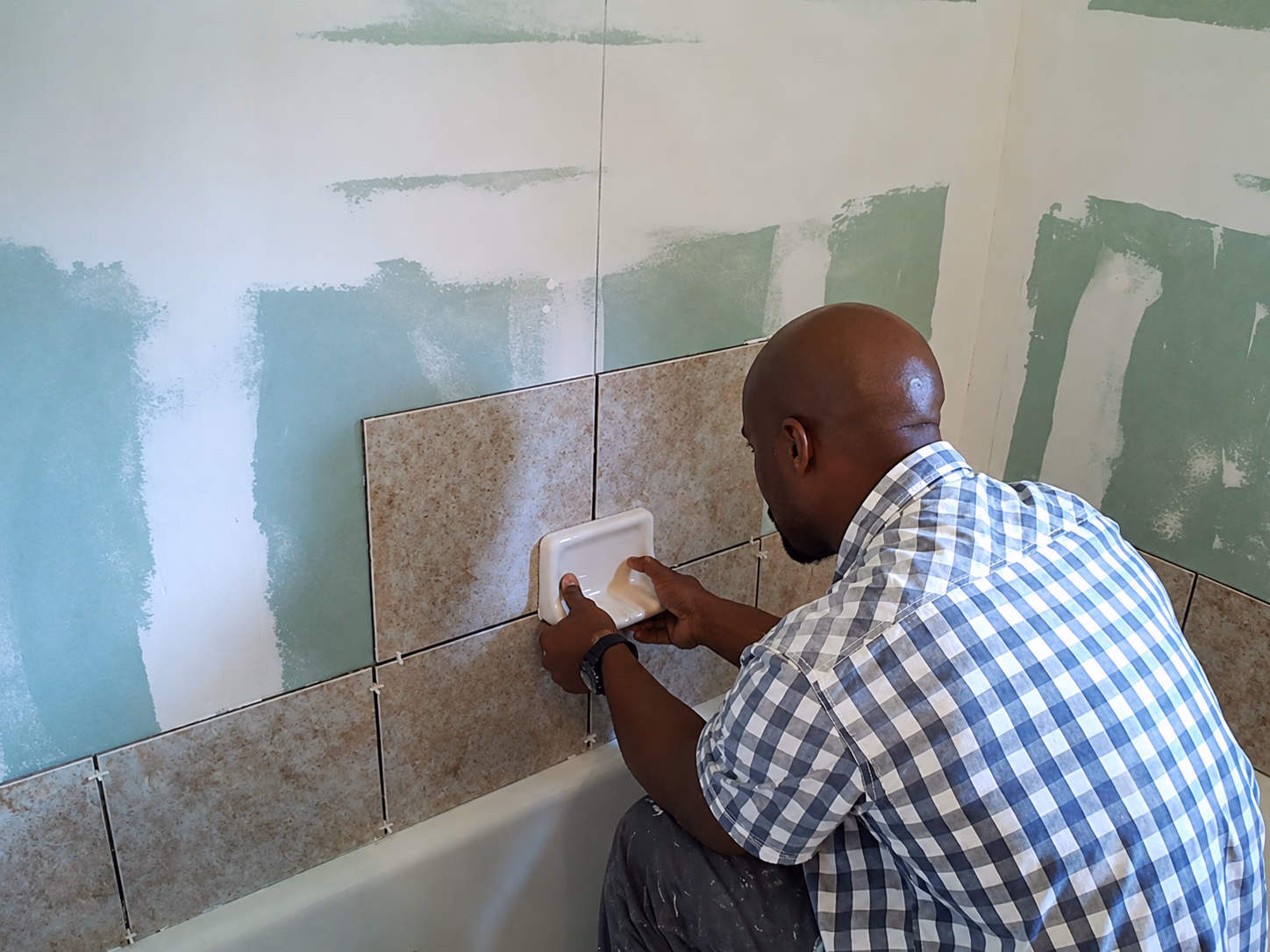 Man in checkered shirt installing wall tiles in bathroom using grout float tool.