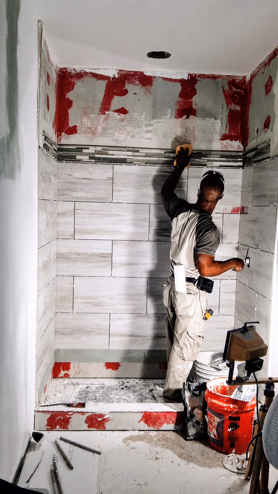 Worker installing beige tiles on shower walls with tools and supplies around him.