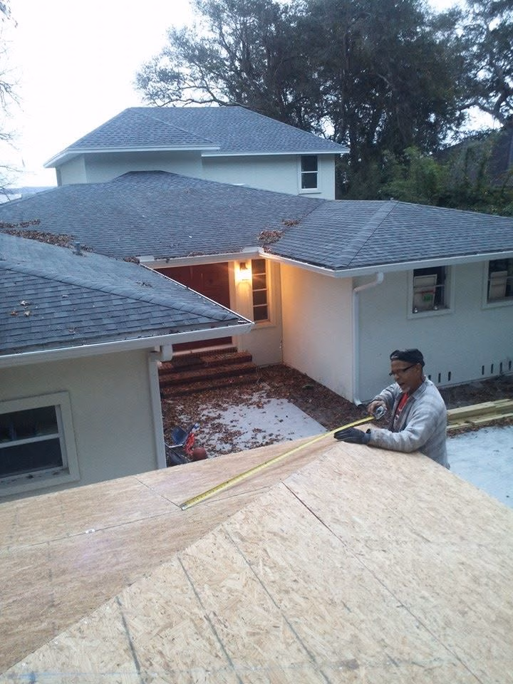 Man measuring wooden roof panels with tape measure in front of white residential house.