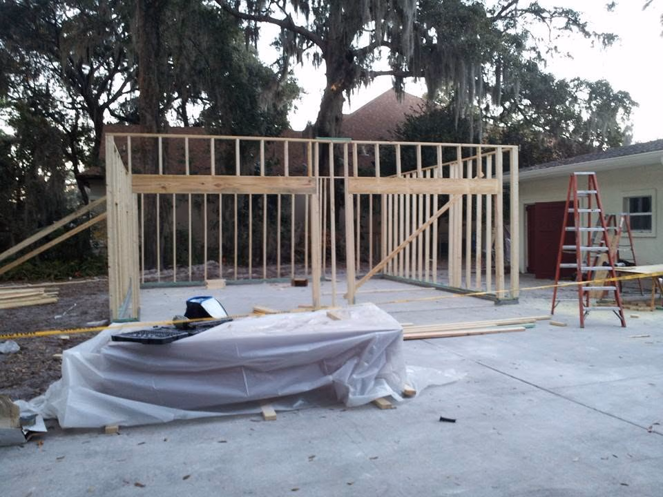 Wooden frame of a small building under construction on a concrete slab with tools and ladders nearby.