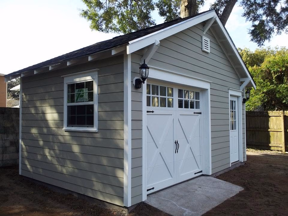 Small gray shed with white trim, double garage door, side window, and entry door under trees.