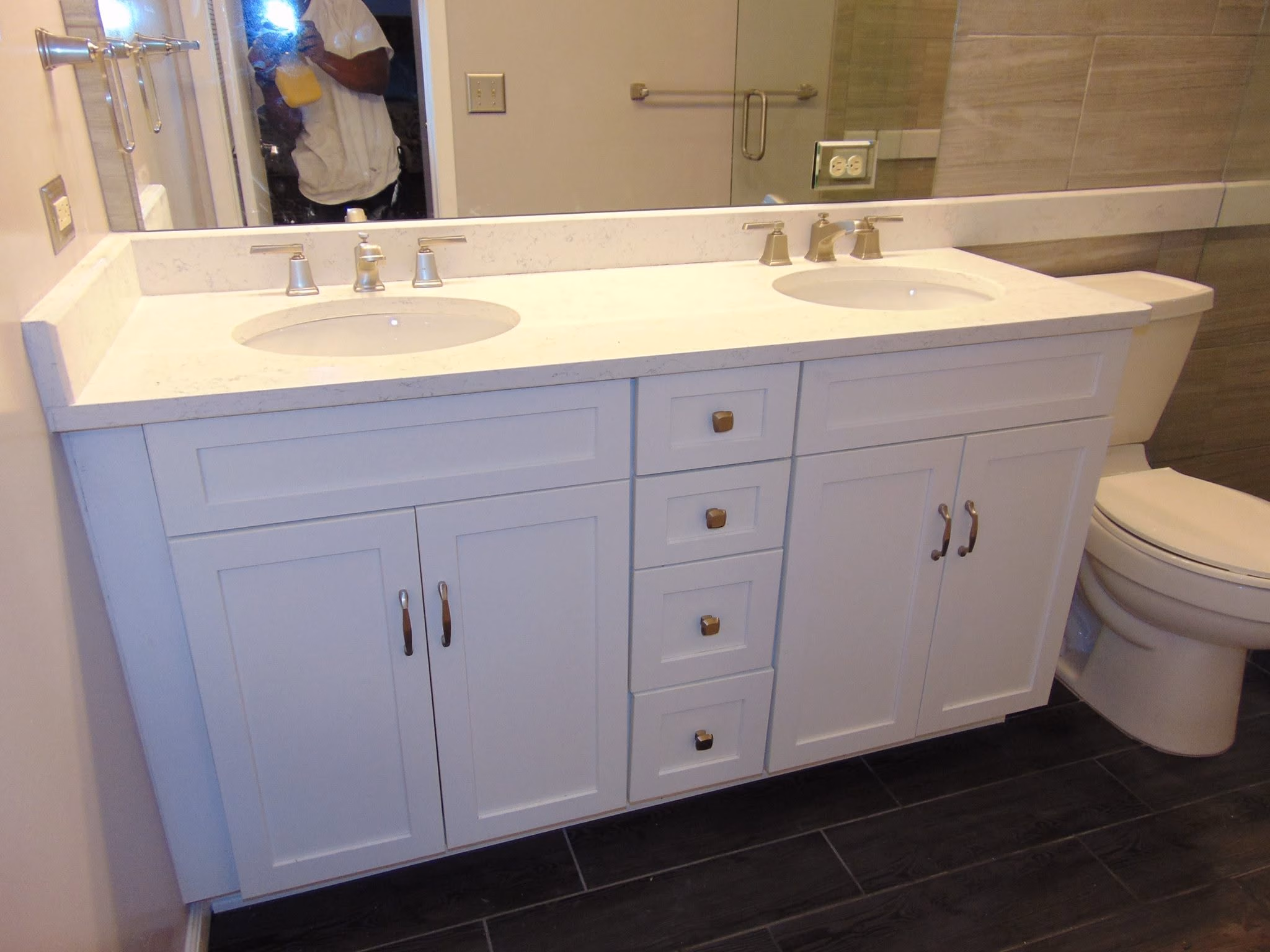Bathroom vanity with double sinks, white cabinets, silver faucets, and a toilet on dark tile floor.