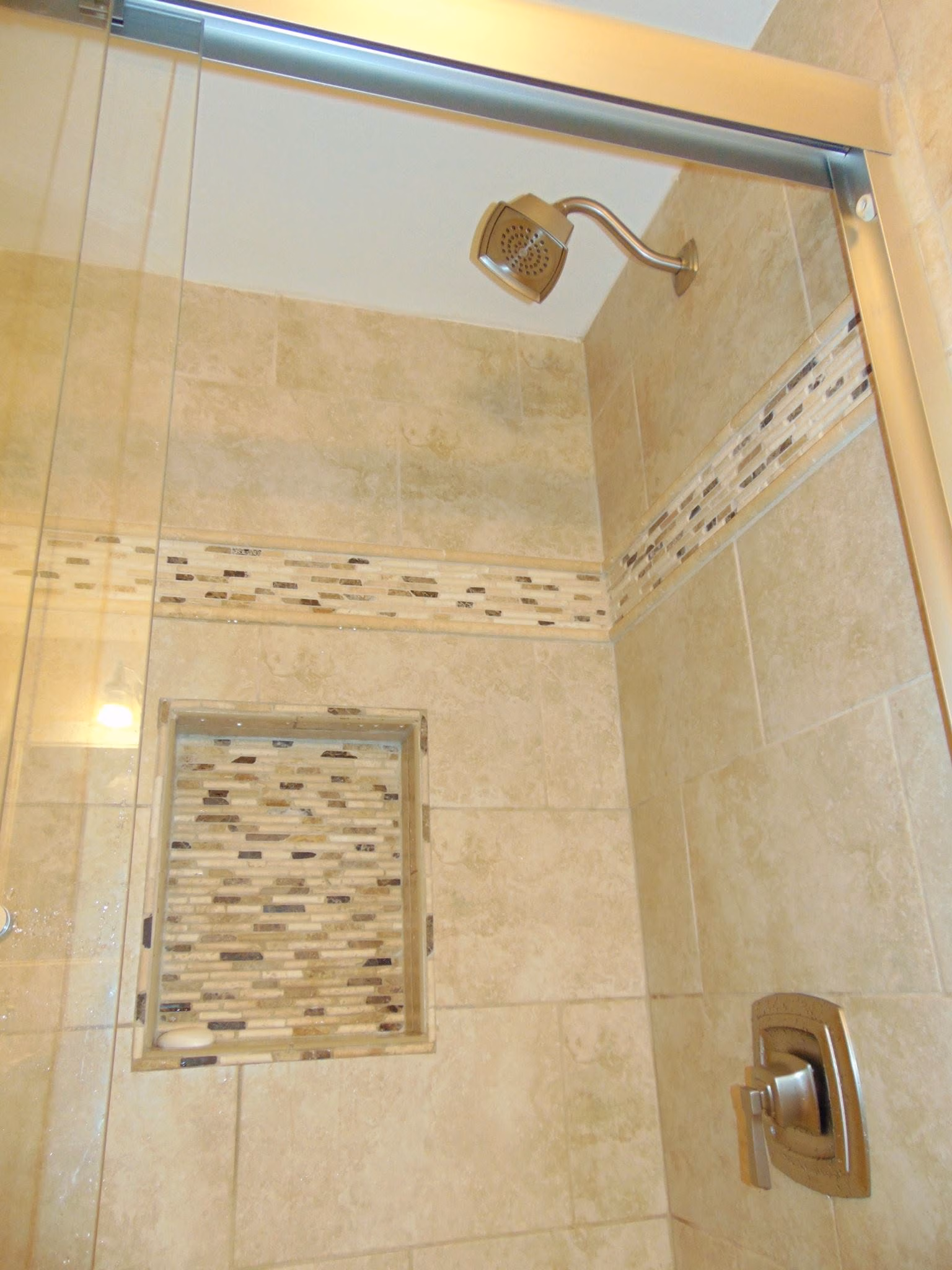 Shower with beige tile walls, mosaic tile accent strip, built-in shelf, and metal showerhead and handle.