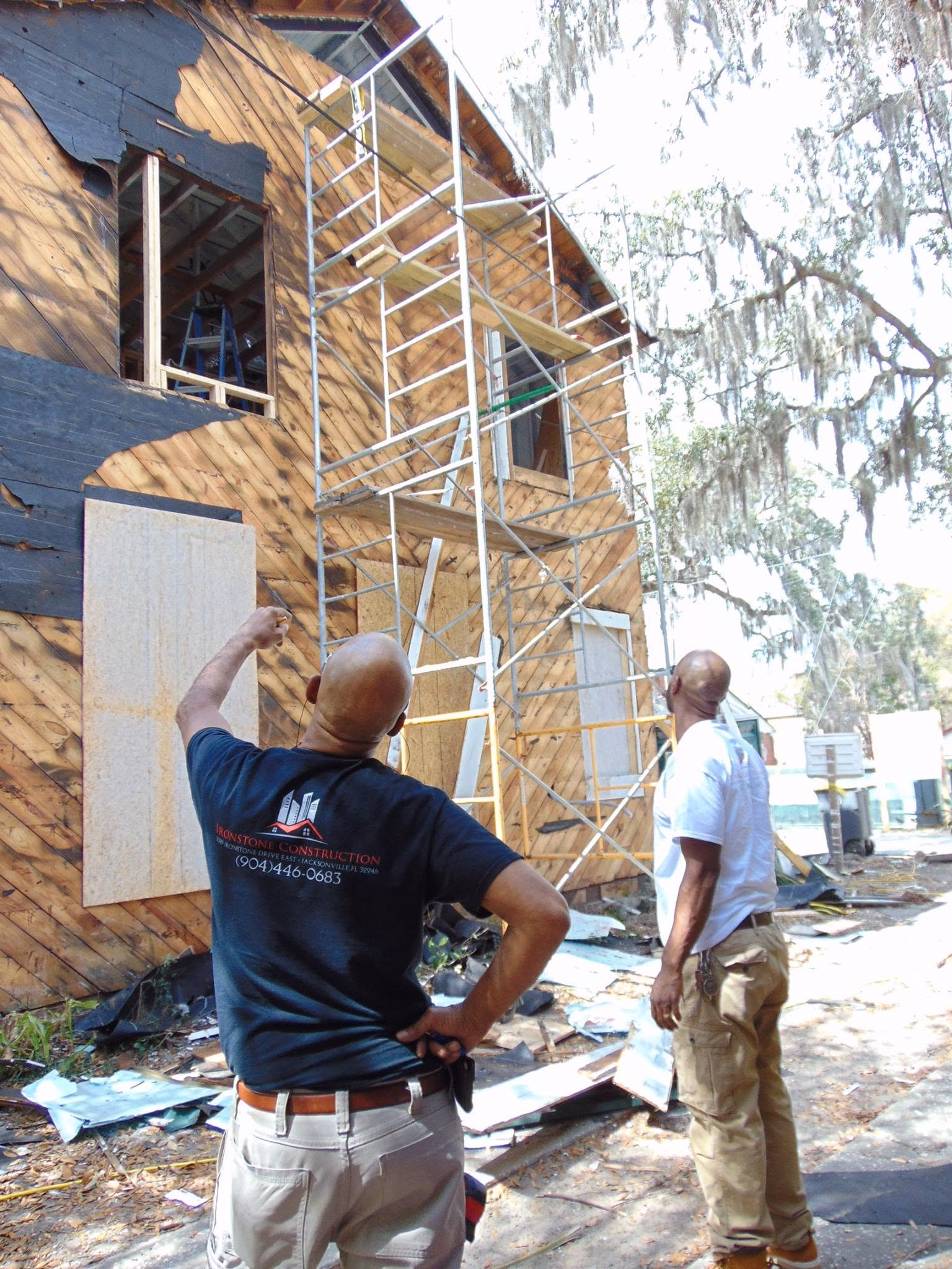 Two men inspecting a wooden house under construction with scaffolding and boarded windows.