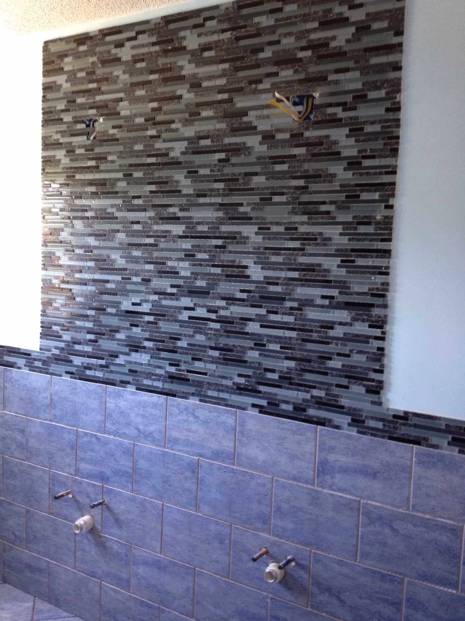 Bathroom wall with upper mosaic tiles in gray tones and lower larger blue tiles, with plumbing fixtures.