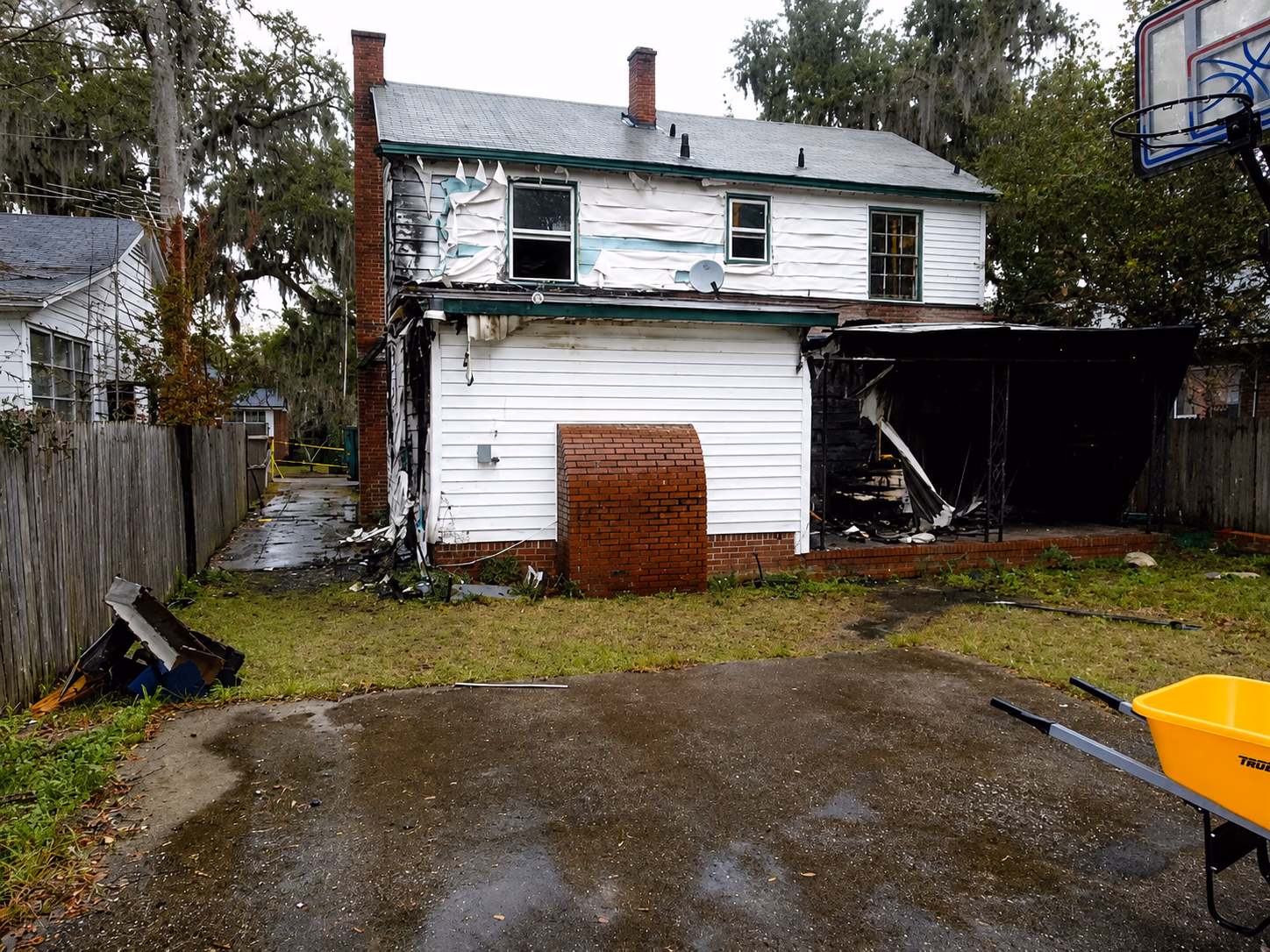 Back of a two-story house showing fire damage with charred walls and peeling siding in yard.