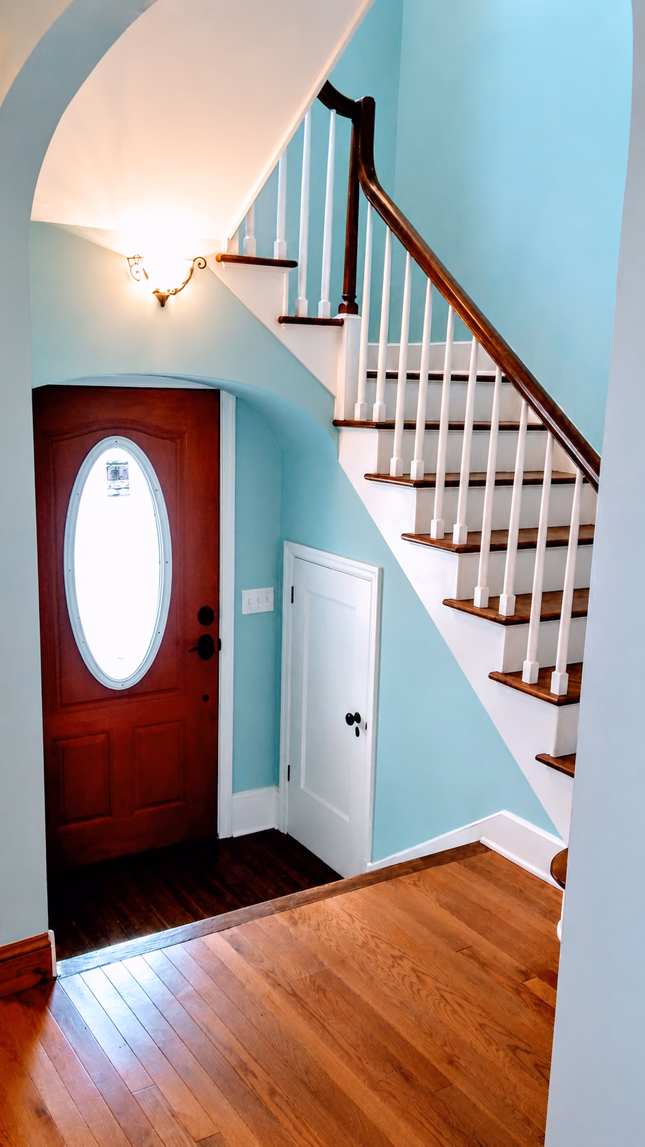 Interior view of a wooden front door with an oval window next to a white staircase with wooden handrails.