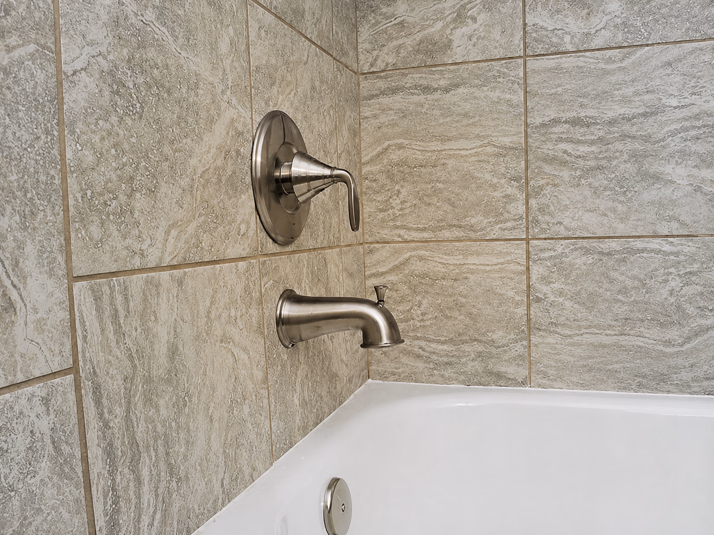 Bathtub faucet and handle mounted on beige marble tile wall above a white tub.