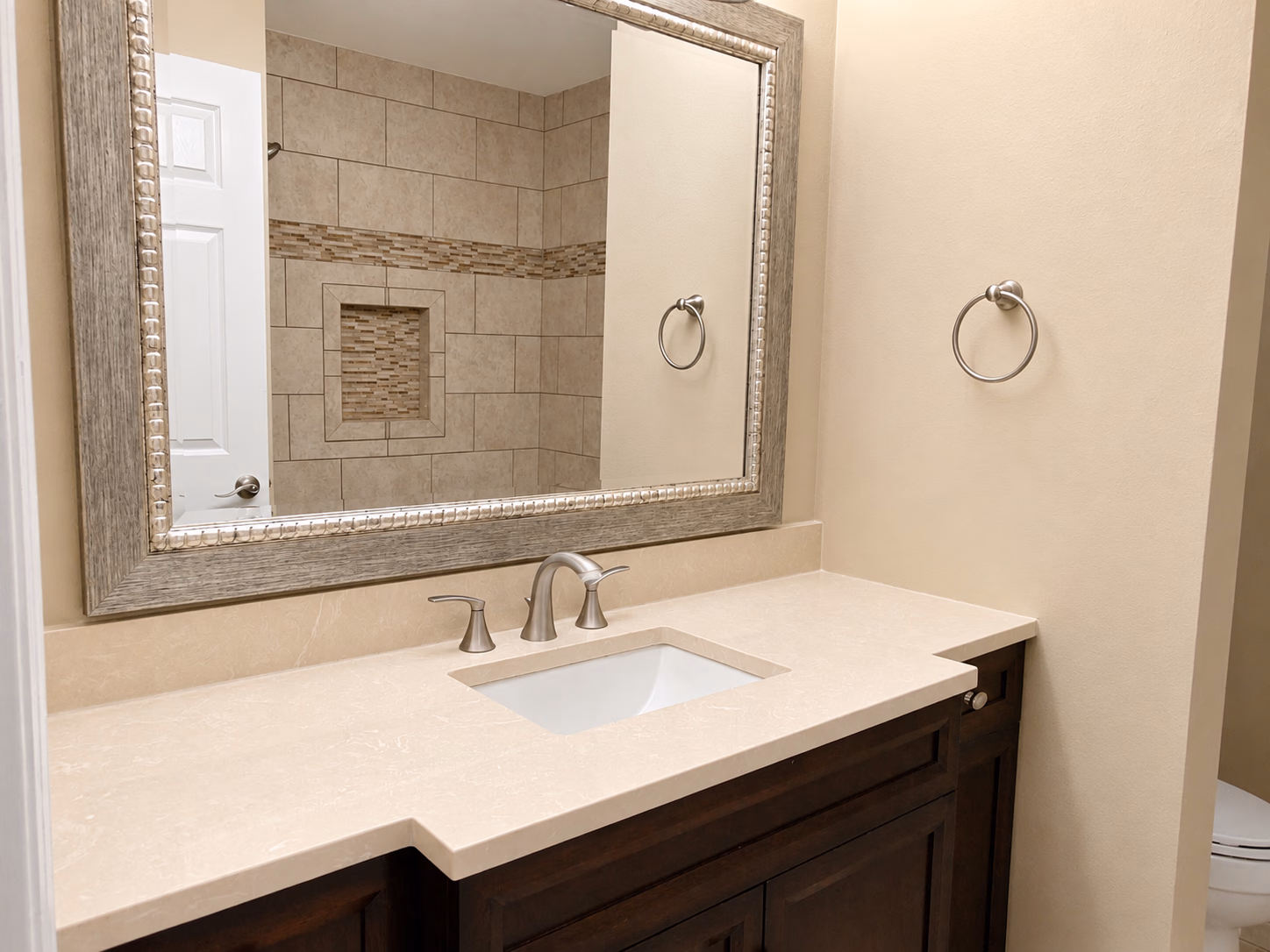 Bathroom sink with beige countertop, silver faucet, large framed mirror, and beige tiled shower reflection.
