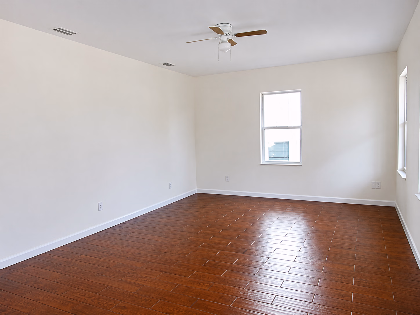 Empty room with white walls, wooden floor, ceiling fan, and two windows letting in daylight.
