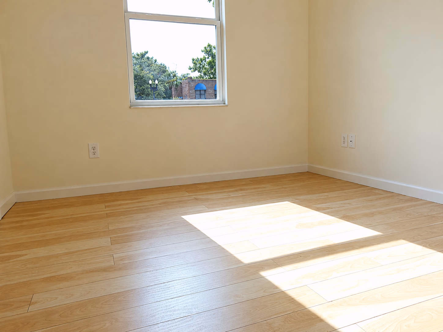 Empty room with light wood floor, beige walls, and a window letting sunlight in.