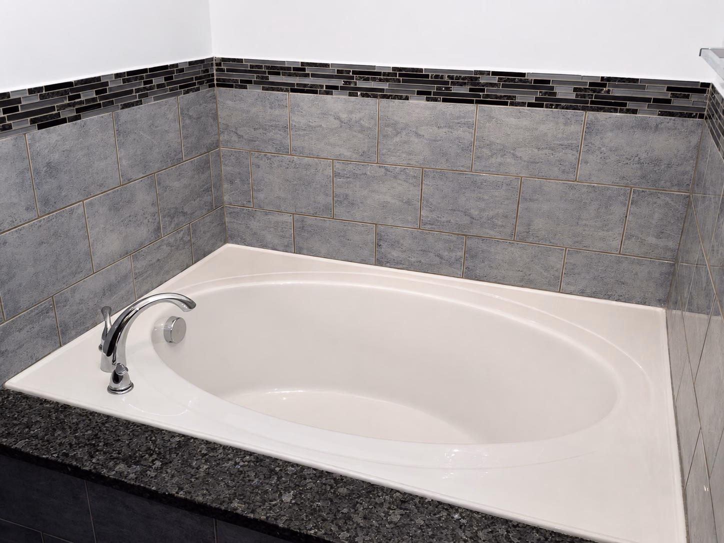 White built-in bathtub with chrome faucet surrounded by gray and black tiled walls and countertop.