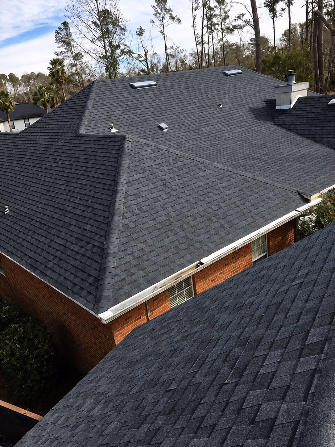 Newly installed dark gray asphalt shingle roof on a brick house with trees in the background.