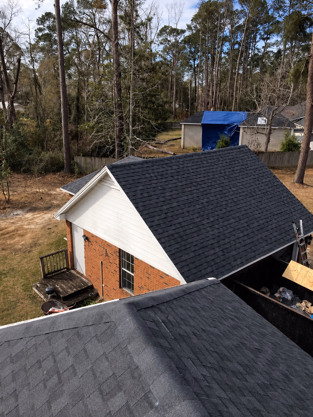 View of dark gray shingled roofs of residential buildings surrounded by tall trees and a yard.
